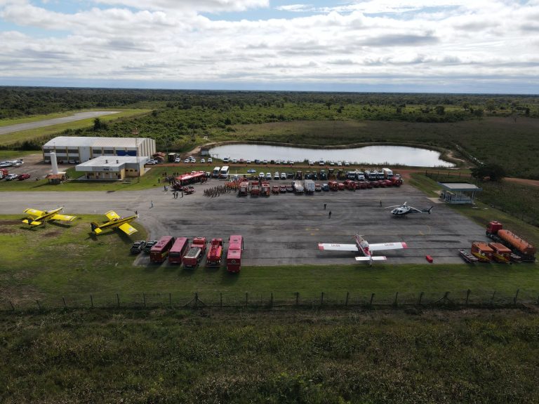 Vista aérea de uma base de combate a incêndios, com aviões, caminhões dos bombeiros e equipes de emergência preparados para atuar.